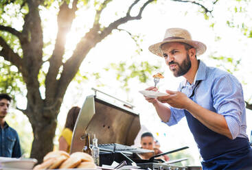 Ein reifer Mann mit Familie und Freunden kocht und serviert Essen auf einer Grillparty im Freien. Familienfeier im Hinterhof. - HPIF28238