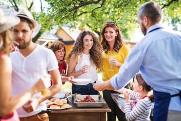 A mature man with family and friends cooking food on a barbecue on a party outside. Family celebration outside in the backyard. - HPIF28242