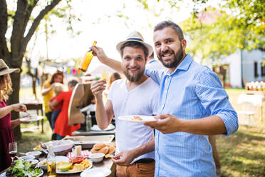 Porträt von Männern, die eine Bierflasche und einen Teller mit Essen in der Hand halten. Eine Familienfeier im Hinterhof. Grillparty. - HPIF28245