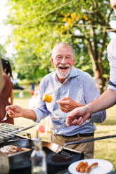 Family celebration outside in the backyard. A happy senior man standing by the grill, holding corn on a barbecue party. - HPIF28248