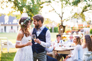 Hochzeitsempfang im Garten, Braut und Bräutigam stoßen mit Champagner an, die Gäste sitzen am Tisch im Hintergrund. - HPIF28255