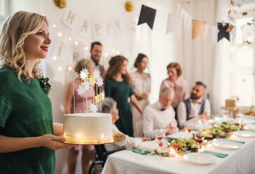 Eine junge Frau hält eine Geburtstagstorte auf einer Indoor-Party, im Hintergrund eine Mehrgenerationenfamilie. - HPIF28299