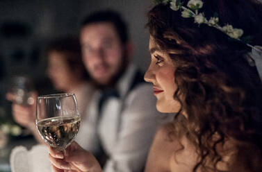 Eine Nahaufnahme einer jungen Braut, die auf einer Hochzeit an einem Tisch sitzt und ein Glas Weißwein in der Hand hält. - HPIF28309