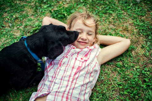 Ein kleines Mädchen mit geschlossenen Augen liegt im Gras, ein schwarzer Hund leckt ihr das Gesicht. - HPIF28317