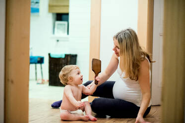 Schöne schwangere Frau sitzt mit einem kleinen Jungen zu Hause auf dem Boden. Ein Kleinkind hält einen Kamm. - HPIF28327