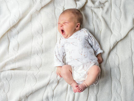 Niedliches, müdes, neugeborenes Baby auf einer weißen Decke liegend, gähnend, Ansicht von oben. - HPIF28329