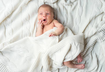 Niedliches neugeborenes Baby mit offenem Mund auf dem Bett liegend, zugedeckt mit einer weißen Decke, Ansicht von oben. - HPIF28331