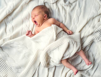 Niedliches neugeborenes Baby auf dem Bett liegend und gähnend, zugedeckt mit einer weißen Decke, Ansicht von oben. - HPIF28332