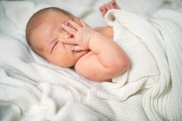 Nahaufnahme eines neugeborenen Babys, das mit einer Hand auf dem Gesicht in einem Bett liegt, zugedeckt von einer weißen Decke. - HPIF28334