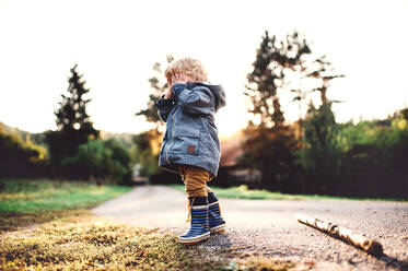 A little toddler boy standing outdoors on a road at sunset, covering his eyes. Copy space. - HPIF28353