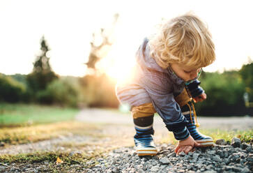 Ein kleiner Junge im Kleinkindalter, der sich bückt und Steine in der Natur bei Sonnenuntergang aufhebt. copy space. - HPIF28357