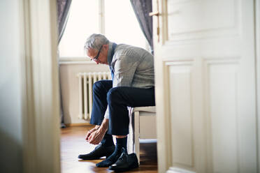 Älterer Geschäftsmann mit Brille auf Geschäftsreise sitzt in einem Hotelzimmer und bindet sich die Schnürsenkel. - HPIF28365