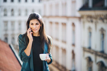 Junge Geschäftsfrau mit Kaffee und Smartphone, die auf einer Terrasse vor einem Büro in der Stadt steht und telefoniert. Kopierraum. - HPIF28374