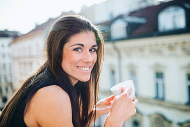 Happy young businesswoman with cup of coffee standing on a terrace in an office in city. - HPIF28383