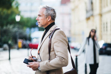 Mature businessman standing on a street in city, holding glasses. Copy space. - HPIF28399