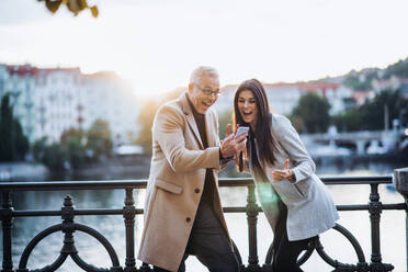 Mann und Frau Geschäftspartner stehen an einem Fluss in der Stadt bei Sonnenuntergang, schauen auf ihr Smartphone und drücken ihre Begeisterung aus. - HPIF28416