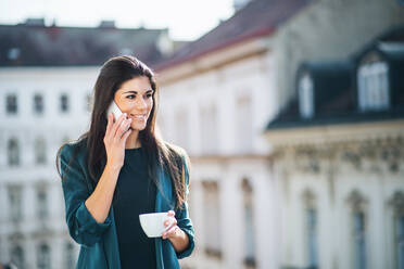 Junge Geschäftsfrau mit Kaffee und Smartphone, die auf einer Terrasse vor einem Büro in der Stadt steht und telefoniert. Kopierraum. - HPIF28432