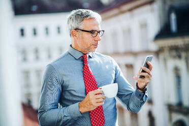 Ein reifer Geschäftsmann mit Kaffee und Smartphone steht auf einer Terrasse in einem Büro in der Stadt und schreibt eine SMS. - HPIF28437