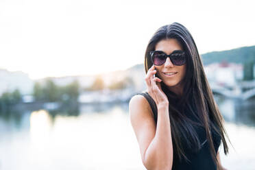 Schöne Frau in schwarzem Kleid mit Sonnenbrille und Smartphone steht an einem Fluss in der Stadt Prag und führt ein Telefongespräch. - HPIF28454