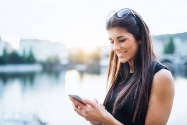 Schöne Frau in schwarzem Kleid mit Sonnenbrille und Smartphone steht an einem Fluss in der Stadt Prag, Text-Messaging. - HPIF28455