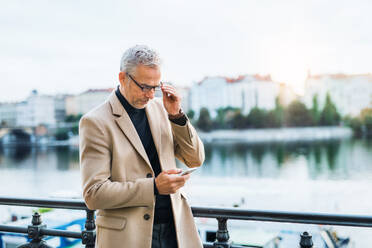 Reifer, gut aussehender Geschäftsmann mit Smartphone, der am Fluss Vltava in Prag bei Sonnenuntergang steht und ein Smartphone benutzt. - HPIF28457