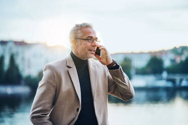 Älterer Geschäftsmann mit Smartphone steht am Fluss in Prag bei Sonnenuntergang und führt ein Telefonat. - HPIF28462