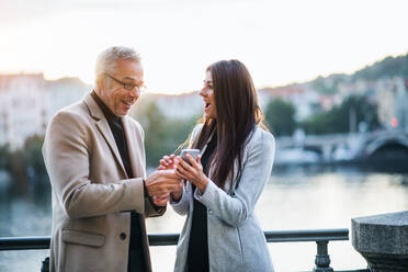 Mann und Frau Geschäftspartner stehen an einem Fluss in der Stadt bei Sonnenuntergang, schauen auf ihr Smartphone und drücken ihre Begeisterung aus. - HPIF28463