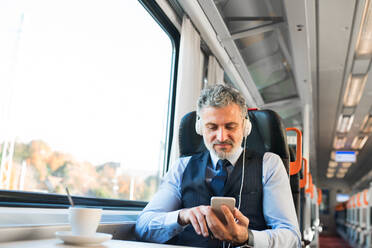 Handsome mature businessman travelling by train. A man with smartphone and headphones, listening to music. - HPIF28478