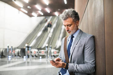 Handsome mature businessman in a city. Man with smartphone standing in front of an escalators at the subway station, text messaging. - HPIF28483