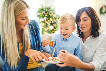 Ein kleiner Junge mit Mutter und Großmutter sitzt zur Weihnachtszeit zu Hause auf dem Sofa und isst Kekse. - HPIF28558