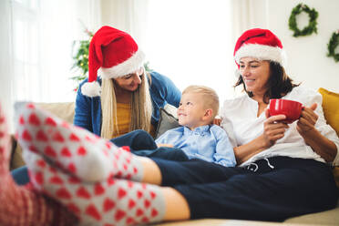 Ein kleiner Junge mit Mutter und Großmutter mit Weihnachtsmannmütze sitzt zur Weihnachtszeit zu Hause auf einem Sofa und unterhält sich. - HPIF28566