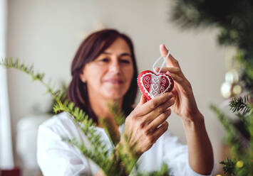 Eine ältere Frau hält ein herzförmiges Ornament in der Hand und schmückt den Weihnachtsbaum zu Hause. - HPIF28569