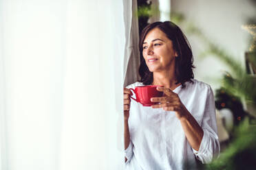 Eine ältere Frau mit einer Tasse, die zur Weihnachtszeit zu Hause am Fenster steht und hinausschaut. Raum kopieren. - HPIF28571