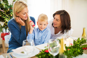 Ein kleiner Junge mit Geschenk und Mutter und Großmutter, die zur Weihnachtszeit zu Hause an einem Tisch sitzen. - HPIF28585