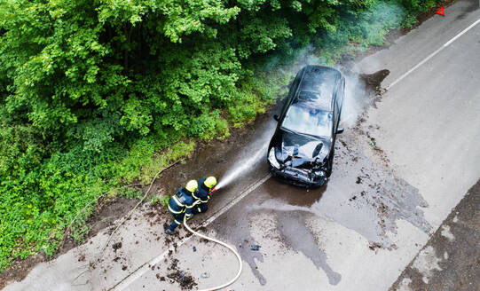 Luftaufnahme von zwei Feuerwehrleuten, die ein brennendes Auto nach einem Unfall auf einer Landstraße löschen. - HPIF28600