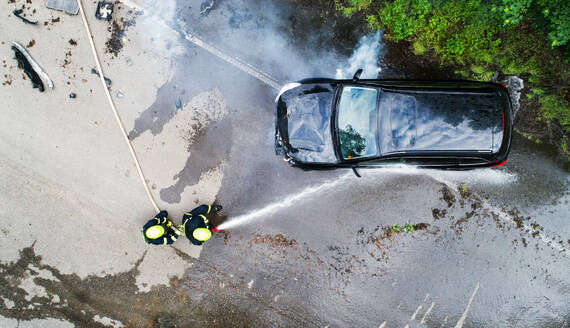 Zwei Feuerwehrleute löschen ein brennendes Auto nach einem Unfall auf der Landstraße, Ansicht von oben. - HPIF28601