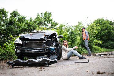 Young frustrated woman sitting by the car after an accident and a man with smartphone, making a phone call. - HPIF28608