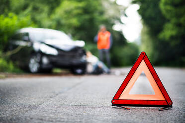 A close up of a red emergency triangle on the road in front of a damaged car and unrecognizable people. A car accident concept. Copy space. - HPIF28609