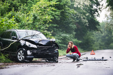 A frustrated young woman with smartphone by the damaged car after a car accident, making a phone call. Copy space. - HPIF28619
