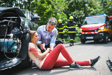 A man helping a young woman on the road after a car accident, firefighters in the background. - HPIF28649