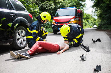 Firefighters helping a young woman after a car accident. A female driver lying down on the countryside road. - HPIF28653