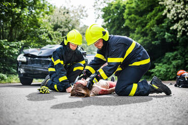 Firefighters helping a young woman after a car accident. A female driver lying down on the countryside road. - HPIF28654