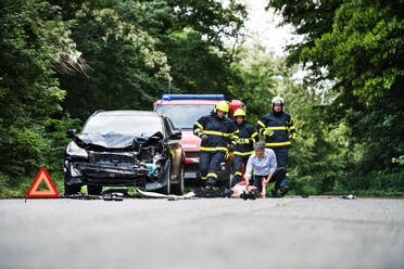 Firefighters running to rescue a woman lying unconscious on the road after a car accident. A mature man holding her hand, checking her pulse. Copy space. - HPIF28656