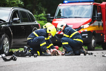 Firefighters helping a young woman after a car accident. A female driver lying down on the countryside road. - HPIF28658