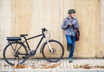 Active senior man with electrobike standing outdoors in town, leaning against a concrete wall and using smartphone. Copy space. - HPIF28680