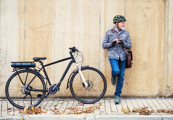 Aktiver älterer Mann mit Elektrofahrrad, der im Freien in der Stadt steht, sich an eine Betonwand lehnt und sein Smartphone benutzt. Raum kopieren. - HPIF28680