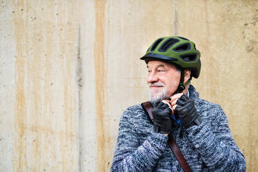 Aktiver älterer Mann, der im Freien an einer Betonmauer in der Stadt steht und einen Fahrradhelm aufsetzt. Raum kopieren. - HPIF28681