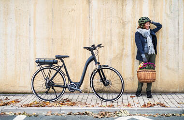 Active senior woman with electrobike and flowers in basket standing outdoors in town, leaning against a concrete wall. Copy space. - HPIF28684