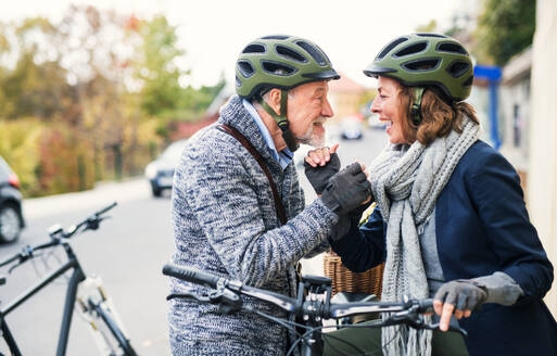 Verliebtes, aktives Seniorenpaar mit Helm und Elektrofahrrad, das auf einer Straße in der Stadt im Freien steht und sich ansieht. - HPIF28687