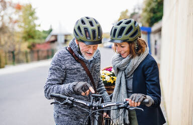 An active senior couple with helmets and electrobikes standing outdoors on a pathway in town, looking at speedometer. - HPIF28688
